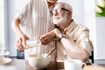 Selective focus of senior man embracing wife pouring milk in bowl during breakfast