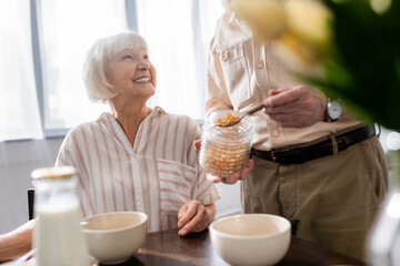 Selective focus of senior woman smiling at husband pouring cereals from jar during breakfast