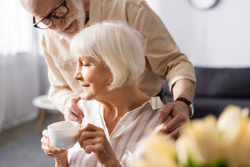 Selective focus of senor man hugging wife with cup of coffee during breakfast at home