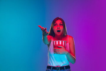 Astonished, eating popcorn. Caucasian young woman's portrait on gradient background in neon light. Beautiful female model. Concept of human emotions, facial expression, sales, ad, movie, cinema.