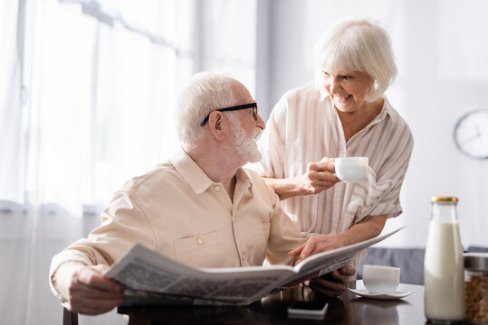 Selective focus of elderly man holding newspaper and smiling at wife with cup of coffee during breakfast in kitchen