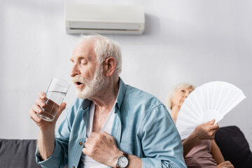 Selective focus of senior man holding glass of water near wife with fan on couch