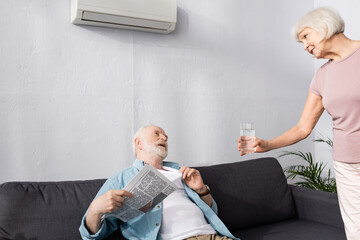 Elderly woman giving glass of water to husband holding newspaper under air conditioner at home