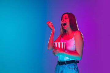 Laughting, eating popcorn. Caucasian young woman's portrait on gradient background in neon light. Beautiful female model. Concept of human emotions, facial expression, sales, ad, movie, cinema.