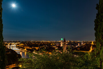 Fototapeta na wymiar Night photo with moon in sky view along Adige river with view of Ponte Nuovo and Ponte Navi with church of Santa Anastasia and Torre di Lamberti, city of Verona, Italy.