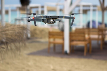 Focus on the modern small folding gray drone. A drone flies and shoots on the seashore and a beach bar can be seen behind.
