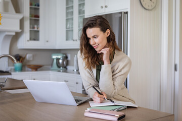 Woman taking notes while using laptop in the kitchen