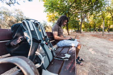 Fototapeta na wymiar Young man with face mask uses laptop alone while keeping social distance to prevent infectious diseases such as coronavirus in the shade of a park with trees in Spain. Selective focus.