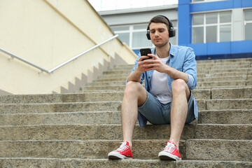 Young man in headphones sits on the steps and looking in phone