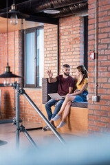 selective focus of businesswoman looking at bearded businessman gesturing near laptop while sitting on window bench in office