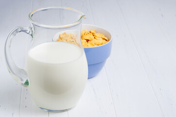 Corn flakes and milk in a glass jug. White wooden table. Copy space