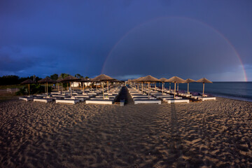 The dark blue sky is above the beach by the sea. It was as if there was a storm, and it turned out to be a rainbow. The beach is empty and there are no people.