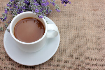 Cup of coffee with milk and lavender flowers on burlap fabric texture with copy space. Enjoying summer morning coffee. Top view. Selective focus