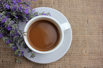 Cup of coffee with milk and lavender flowers on burlap fabric texture with copy space. Enjoying summer morning coffee. Top view. Selective focus