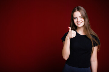 Beautiful long-haired girl in a black T-shirt shows a thumb as a sign of approval and like on a red background