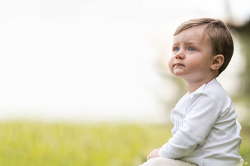 1 year old caucasian boy sitting on green lawn, with white clothes