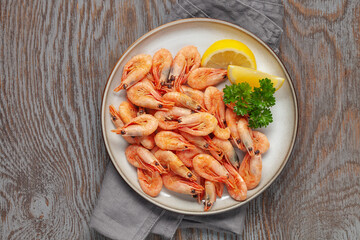 Boiled shrimp prawns in a shell with lemon and parsley in a plate on a wooden background. Top view.