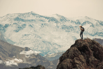 Mujer haciendo senderismo parada en la cima de una montaña muy alta. Mira hacia abajo se ve un valle y nubes, de fondo la cordillera - obrazy, fototapety, plakaty