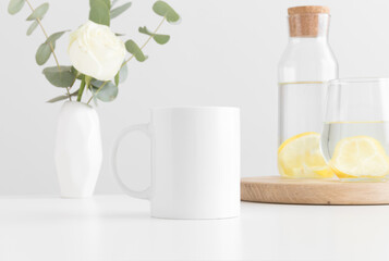 White mug mockup with flowers in a vase, glass and a bottle on a white table.
