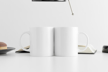 Two white mugs mockup with a donut, book and a lamp on a white table.
