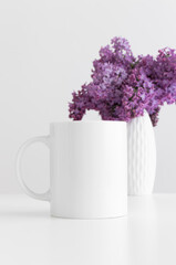 White mug mockup with a bouquet of lilac in a vase on a white table.