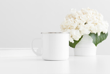 Enamel mug mockup with a book and a hortensia in a pot on a white table.
