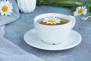 Herbal chamomile tea with fresh chamomile flowers in a white cup and saucer on a gray background. Bouquet of flowers on a wooden board and linen napkin.