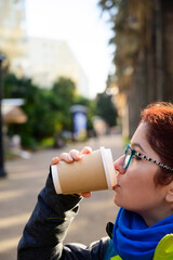 Red-haired caucasian woman in glasses and a jacket is drinking coffee on the street. Girl enjoys a warming drink on a cold winter day.