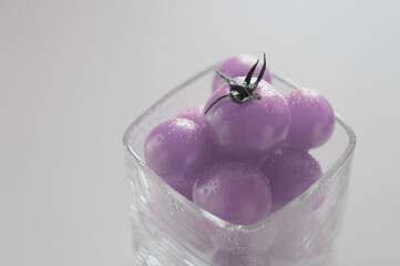 Lilac cherry tomatoes in a square glass with drops of water on a gray background. Creative horizontal photo, pastel colors with place for text.