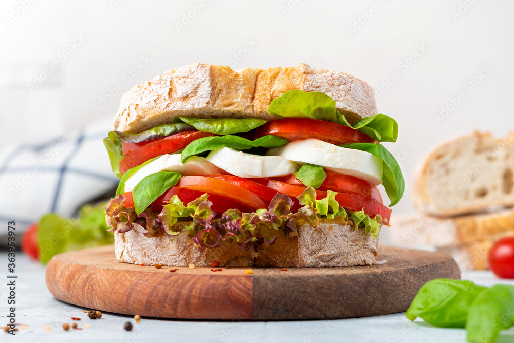 Sticker Big tasty sandwich of ciabatta with mozzarella cheese, tomatoes, basil and lettuce on the table. Sandwich with cheese and vegetables on a light background, vegetarian food. Italian Cuisine.