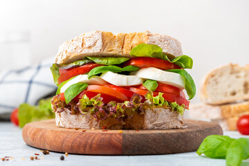 Big tasty sandwich of ciabatta with mozzarella cheese, tomatoes, basil and lettuce on the table. Sandwich with cheese and vegetables on a light background, vegetarian food. Italian Cuisine.