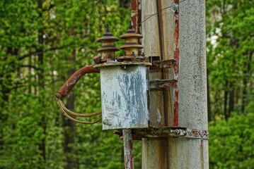gray iron transformer in brown rust with wires on a concrete pole against a background of green vegetation