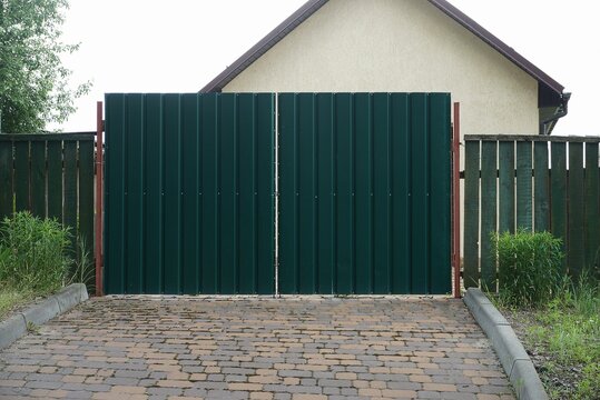 large closed iron green gate and part of a wooden fence on a brown sidewalk on a rural street