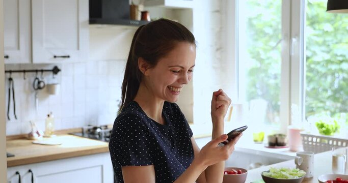 Woman spend time in kitchen distracted from cooking vegetable salad dinner preparation holding smart phone read great news. Moment of sincere happy emotions, win, victory opportunity, new app concept