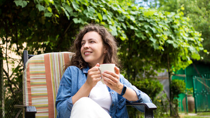 Portrait of dreamy young woman enjoying drinking coffee in the morning sitting on chair on home terrace on green garden background.Happy morning routine,day planning,enjoying life,to be in the moment