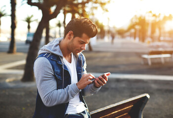 Portrait of handsome young man sending a text message while standing outside, male student reading text messages outdoors, stylish brunette hipster using cell phone at sunny evening, flare sunshine