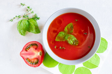 Top view of cold gazpacho soup in a grey bowl with half of a tomato and herbs on a light grey background