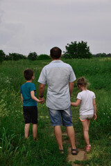 A friendly family, father holds the hand of a son and daughter, they walk together in a green meadow. A boy and a girl with his father go along the road and go into the distance.