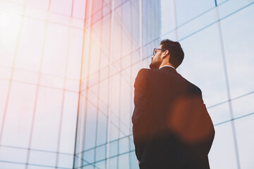 Back view of serious businessman in eye glasses looking on copy space while standing against glass skyscraper, young professional employee waiting for international partners outdoors near big company