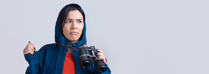 A tourist girl in a blue raincoat holds binoculars in her hands and looks into the distance, spies.