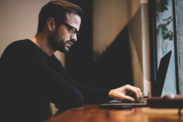 Serious concentrated designer typing text message on keyboard of laptop searching important information using internet sources and working remote in cozy contemporary interior at wooden table