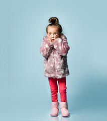 Little blonde kid with bun hairstyle, dressed in pink faux fur coat. She is looking excitedly, posing on blue background. Close up