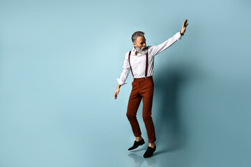 Man in years in white shirt, brown pants, black loafers. He runs looking back, showing stop by his hand, posing on blue background