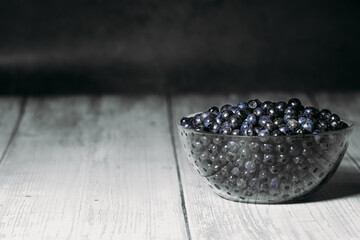 A transparent glass plate is filled with ripe tasty blueberries on a dark background. Copy space. Place for text