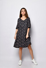 Brunette model in black dress with floral print, white sneakers. Smiling, touching her face, posing isolated on white. Full length