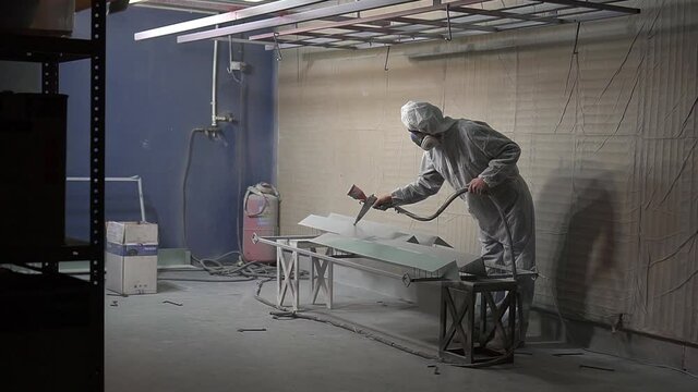 Powder coating of metal products. A man in a White protective suit and a respirator with a spray gun applies a gray powder to an object in the paint chamber.