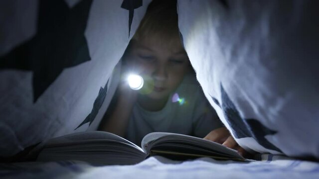 A little boy reads a book with a flashlight under the covers at night.