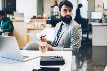 Creative manager of corporation working on solutions for business project sitting at laptop device in office and making notes while looking away.Bearded banker in formal wear fills in questionnaire