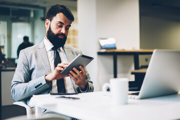 Bearded male banker in formal apparel reading last financial news on modern touch pad device connecting to 4G internet sitting in office at workplace with digital computer during recreation time