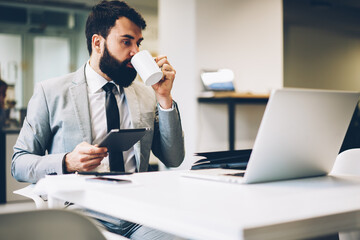 Fototapeta premium Bearded executive manager with digital tablet in hand drinking tasty coffee while watching online broadcast with latest business events on modern laptop device connected to wireless internet in office